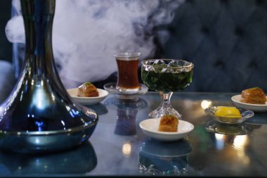 Bir nargile tabanı çay, yeşil bir içecek ve baklava ve limon dilimli birkaç küçük tabak ile bir masada oturur. Duman arka planı doldurur.