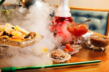 Çeşitli atıştırmalıklar ve nargile piposu olan bir masa, pipodan yükselen bir duman ve yakınlardaki bir içecek..