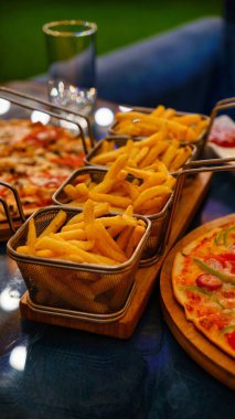 Altın patates kızartmasıyla dolu üç tel sepet bir masada, arka planda bir pizzayla oturuyor..