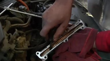Bir tamirci elinde metalik bir motor parçası tutuyor, yerine oturtmadan önce kontrol ediyor. Rol yağ ve kir içinde, aşınma ve yırtık izleri gösteriyor..