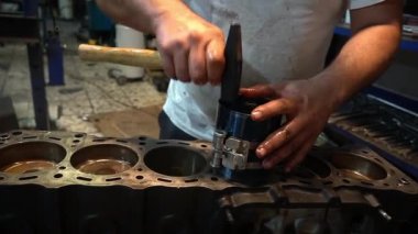 Bir tamirci motor parçası takmak için çekiç kullanıyor. Parça silindir bir kafaya yerleştiriliyor ve tamirci eldiven takıyor..