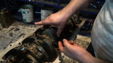 Bir tamirci araba motoru üzerinde çalışır, parçaları inceler ve ayarlar. Motor bloğu açık, krank mili ve diğer bileşenleri ortaya çıkarıyor.