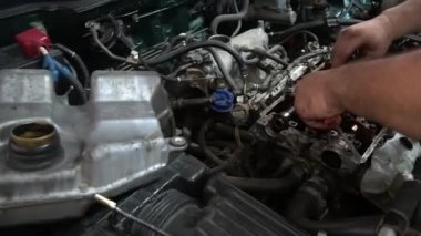 Bir tamirci bir arabanın motoru üzerinde çalışıyor, muhtemelen parçaları değiştiriyor ya da ayarlıyor. Motor bölmesi açık, çeşitli hortum ve bileşenleri ortaya çıkarıyor..