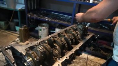 Bir tamirci garajda bir araba motoru üzerinde çalışıyor, sürgüyü sıkıştırmak için İngiliz anahtarı kullanıyor. Motor açık, krank mili ve diğer bileşenleri ortaya çıkarıyor.