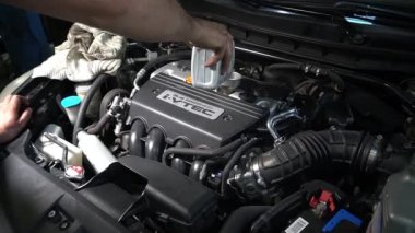 Bir tamirci arabanın motoruna yağ döküyor. Arabanın kaputu açık ve motor görünür durumda..