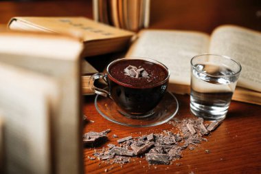 Çikolata parçacıklı bir fincan sıcak çikolata, açık bir kitap ve bir bardak suyun yanındaki ahşap masaya oturur..