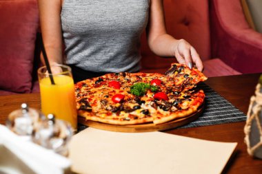 Restoranda bir kadın bir dilim pizza alıyor, masada bir bardak portakal suyu, tuz ve biberlikle..