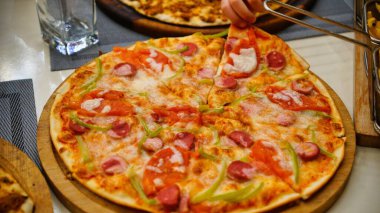 Bir el, ahşap tabakta servis edilen bir dilim pepperoni pizzasına uzanır. Pizzada yeşil biber, domates, sosis ve peynir var..