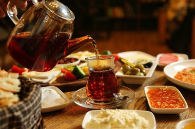 Bir el, zeytin, peynir ve ekmek gibi kahvaltılık gıdalarla çevrili, cam çaydanlıktan geleneksel bir bardağa Türk çayı dolduruyor..