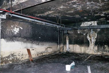 Yangın yüzünden kurum ve su hasarı olan bir bodrum katı. Duvarlar ve tavan kararmış ve lekelenmiş. Yerde beyaz bir kova ve plastik bir çanta var..