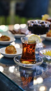 Limon dilimli bir bardak Türk çayı, içinde hamur işleri ve bulanık bir arka plan olan cam bir masanın üzerinde oturuyor..