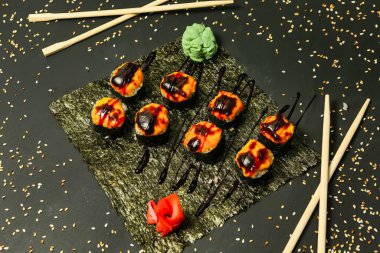 Sushi dürümleri siyah bir tabakta susam tohumlarıyla sıralanmış. Yanında yemek çubukları ve küçük bir parça wasabi var..
