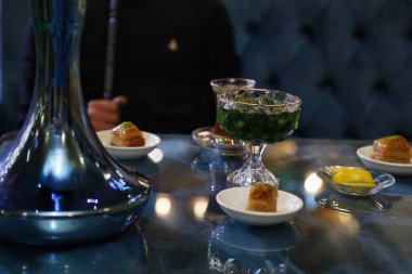 Mavi bir shisha tabanı, yeşil bir içecek ve bir kafede cam bir masada oturan bir tabak baklava..