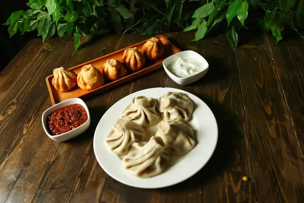 Geleneksel Gürcü hamur tatlısı Khinkali, baharatlı kırmızı ve beyaz soslu, ahşap bir masada dinleniyor. Yakınlarda fazladan köfte tepsisi var..