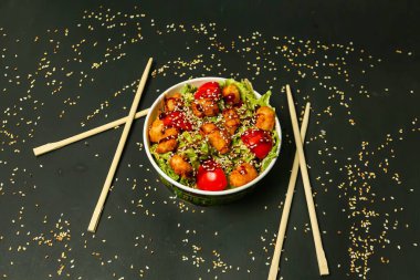 Susam tohumlu bir kase tavuk ve domates salatası yanında iki çift çubukla susam tohumuyla siyah bir zemin üzerine serpiştirilir..