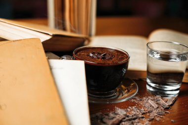 Çikolata parçacıklı bir fincan sıcak çikolata, bir bardak su ve ahşap bir masanın üzerinde bir yığın kitap..