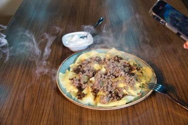 A plate of steaming Turkish food, likely a type of manti or ravioli, topped with ground meat and served with a side of yogurt. clipart