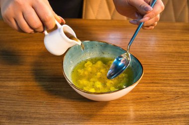 Bir el, küçük beyaz bir sürahiden çorba kasesine çorba döker. Kasede de bir kaşık var..