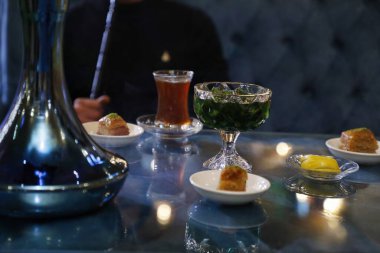 Nargile tabanlı bir masa, çay, yeşil bir içecek ve birkaç küçük tabak baklava..