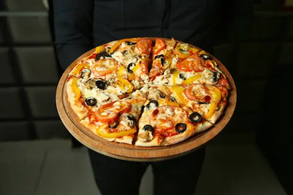 Sarı biberli, domates dilimli ve siyah tişörtlü birinin tuttuğu tahta tahtada siyah zeytinli bir pizza..