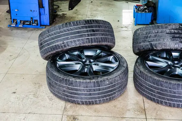 İki yeni siyah araba lastiği ve jantlar garajın zemininde duruyor. Lastikler üst üste yığılmış, siyah alaşımlı jantları gösteriyor..