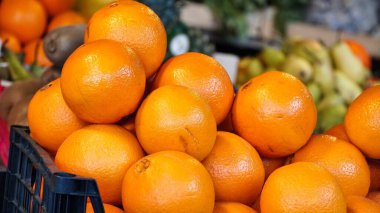 Parlak turuncu portakal portakalları, arka planda odaklanamayan yeşil meyvelerle çevrili siyah plastik bir sandıkta üst üste yığılmış..