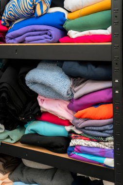 Bir raf ünitesinde farklı renk ve kumaşlarda çeşitli katlanmış giysiler düzgünce düzenlenir. Düzenlemede canlı tekstil ve giyim türleri gösteriliyor.