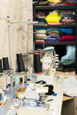 Bir dikiş atölyesinde birden fazla dikiş makinesi ve iplik makarası sergilenirken, raflarda çeşitli renklerde kumaş ruloları kullanıma hazır..