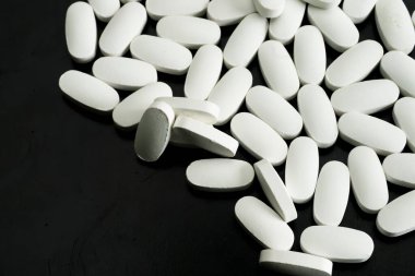 Karanlık bir yüzeye yayılmış sayısız beyaz oval tablet, çeşitli ilaç ürünlerini ve bunların ayırt edici şekillerini vurguluyor..