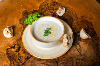 Bir kase kremalı çorba, maydanozla süslenmiş, ahşap bir masanın üzerinde oturur. Etrafı mantarlarla çevrilidir..