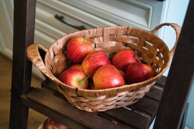 Canlı kırmızı elmalarla dolu dokunmuş bir sepet ahşap bir rafta duruyor. Ev mutfağındaki taze ürünleri ön plana çıkaran ortam rahat ve sade..