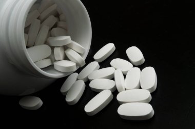 Beyaz kapsüller bir kabın içinden karanlık bir yüzeye dökülür, pürüzsüz dokuları ve düzgün şekilleri sergilenir. Odak karşıtlığı vurgular.