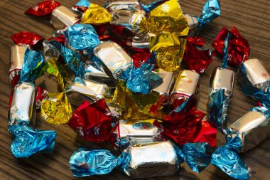 Metalik mavi, kırmızı ve sarı renklere sarılı çeşitli şekerler ahşap bir masaya saçılmış, neşeli bir atmosfer yaratıyor..