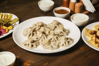 Konforlu bir restoran masasında yoğurt, sos ve turşu tabağının yanında bir tabak hamur tatlısı sunulur. Lezzetli bir yemek deneyimini davet eder..