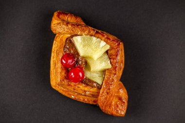 Bu hamur işi, ananas ve kirazla kaplı, koyu bir arka planda sunulmuş, hoş bir tat deneyimini davet eden pullu bir kabuğu içeriyor..