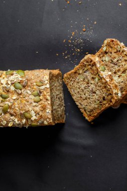 Bir somun tam tahıllı ekmek ve üzerinde çıtır çıtır bir tohum var. Karanlık bir yüzeyde oturuyor. Dilimler kesilmiş, içerideki dokuyu gösteriyor. Pişirme ve sağlıklı beslenme vurgulanır.