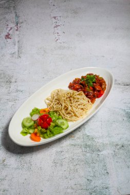 Taze sebze salatası ve baharatlı sosun yanında şehriye içeren canlı bir yemek. Hepsi de minimalist bir arkaplanda, parlak beyaz bir tabakta sunuluyor..