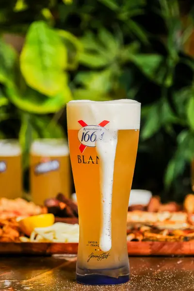 Ağzına kadar dolu buzlu bir bardak bira canlı bir yiyecek serpintisinin önünde gururla durur. Arka planda yeşillik ve daha fazla içki yer alıyor.