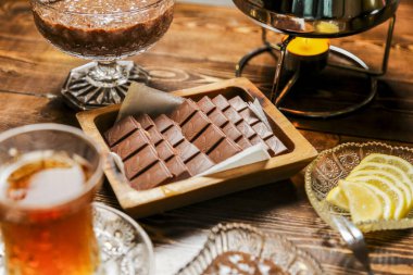 A wooden table is set with an assortment of chocolate squares, a glass of tea, and slices of lemon. The warm glow of a candle adds a cozy atmosphere. clipart