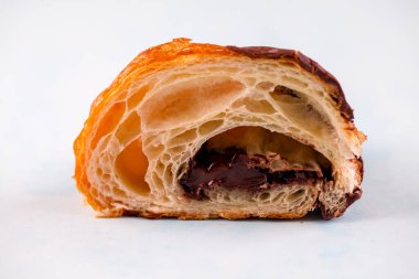 Yeni pişmiş bir kruvasan, pul pul dökülmüş tabakaları ve bozuk çikolata dolgusu ile ikiye bölünür. Tatlı, açık mavi bir arkaplana dayanır..