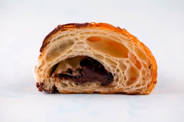 Bir çikolatalı kruvasan ikiye bölünür, yumuşak katmanlarını ve yapışkan çikolata dolgusunu ortaya çıkarır. Arka plan pastayı vurgulayan basit bir açık renk..