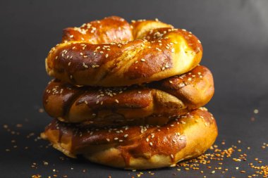 Üç tane çiğnenmiş ve kahverengi ekmek düğümü üst üste istiflenmiş, üzerine susam tohumu serpiştirilmiş, koyu bir arka planda dinleniyor. Doku davetkar ve taze görünüyor..