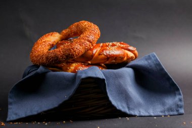 Örgü ve halka şeklinde hamur işleri de dahil olmak üzere çeşitli yeni pişmiş ekmekler sepetteki mavi bir bez üzerinde sunuluyor, altın dokuları ve susam tohumları vurgulanıyor..