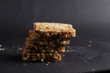 Düzgünce istiflenmiş çoklu tahıllı ekmek dilimleri, koyu bir zemin üzerinde tohum ve tahıl karışımı sergiliyor, sağlıklı dokusunu ve cazibesini vurguluyor..