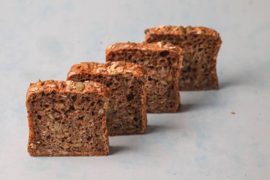 Dört dilim tam tahıllı ekmek açık mavi bir yüzey üzerinde yan yana dizilmiştir. Ekmek tohumlar ve taneler içeriyor, sağlıklı bir doku sergiliyor..