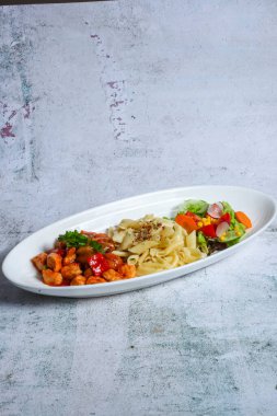 İyi sunulmuş oval tabakta, üzerinde karides ve kırmızı sos bulunan bir porsiyon makarna ve yanında da renkli bir sebze salatası bulunur..