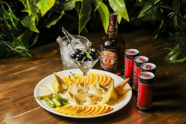 Muz, portakal ve kivi dahil taze kesilmiş meyveler beyaz bir tabakta düzenlenir. Yakınlarda bir şişe viski, mikser ve kokteyl aksesuarları var..