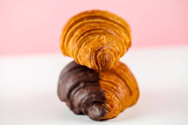İki yeni pişmiş kruvasan üst üste istiflenmiş, tepesinde altın bir hamur işi ve altında pembe bir zemin üzerine boyanmış çikolata kaplı bir hamur işi var..