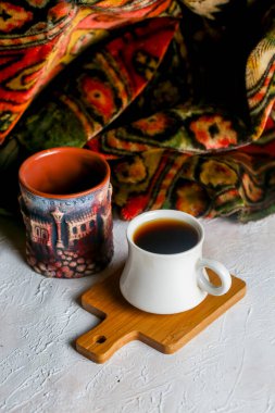Koyu kahve renkli bir seramik fincanın yanında, canlı bir tekstil ile çevrili, beyaz bir fincanda oturur ve ahşap bir tahtanın üzerine konur..