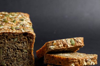 Tohumlu tam tahıllı ekmek dilimleri karanlık bir yüzeyde oturur, kabuklu bir dış görünüş ve yumuşak bir iç görünüş sergilerler. Doku sağlıklı bileşenleri vurguluyor..